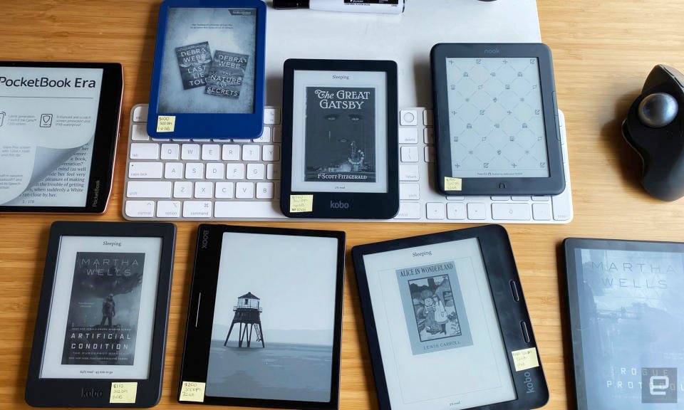 An assortment of ereaders, including ones from Kobo, Kindle, Boox, Nook and PocketBook are arranged on a tan bamboo desk with a white keyboard and black mouse in the background.