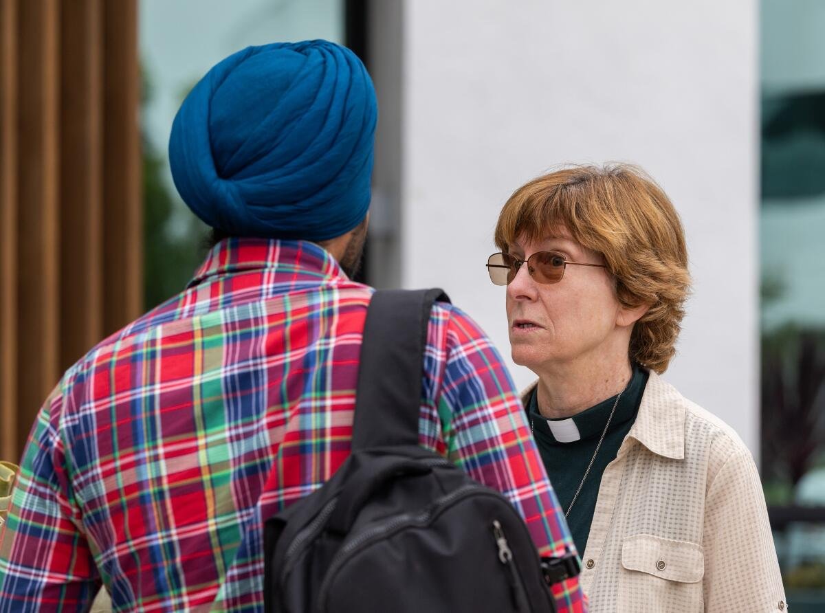 a woman priest speaks to a man with a blue turban and plaid shirt